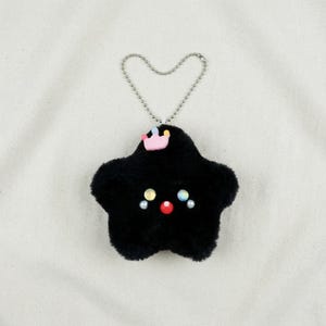 May include: A black star-shaped plush keychain with a small pink crown and colorful facial details. The keychain has a silver chain and a fluffy texture, creating a whimsical and cute design.