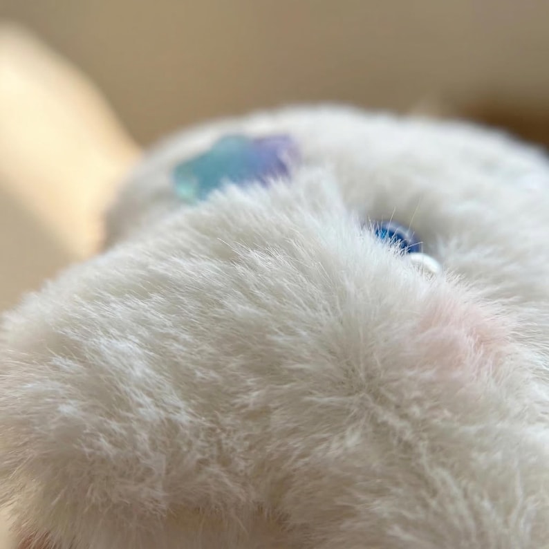 May include: Close-up of a white, fluffy object with a soft texture. A small, iridescent blue and purple gem is visible on the surface. The object has a slight pink hue in one area.