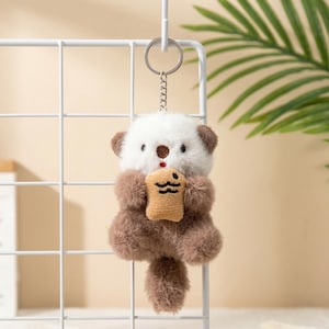 May include: A plush keychain featuring a cute otter with a white head, brown ears and body, holding a small beige snack. The keychain has a silver chain and ring for easy attachment. The otter's face has small black eyes and a brown nose.