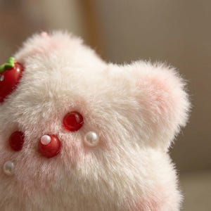 May include: A close-up of a plush, white and pink strawberry-shaped item. The item has a fluffy texture and is adorned with red, round embellishments and white pearls. A small, red strawberry is visible on the top left.