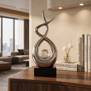 May include: A modern silver sculpture with an abstract design, featuring a central orb. The sculpture sits on a black rectangular base. The background includes books, a vase with flowers, and a wooden surface.