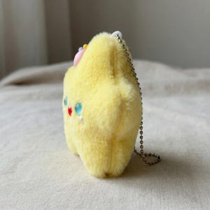 May include: A plush yellow star-shaped keychain with a fluffy texture. It features colorful beaded accents, including red, blue, and white, and a small pink bow. A silver chain is attached for hanging.