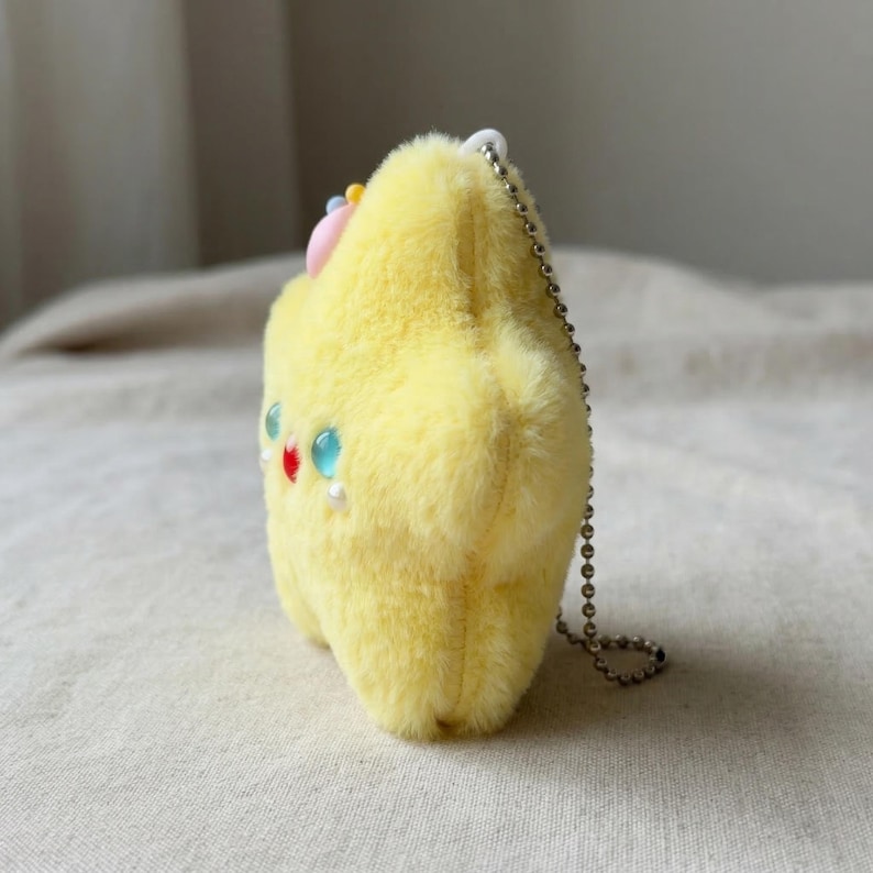 May include: A plush yellow star-shaped keychain with a fluffy texture. It features colorful beaded accents, including red, blue, and white, and a small pink bow. A silver chain is attached for hanging.
