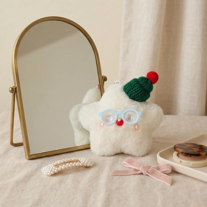 May include: A plush white star-shaped toy with blue glasses, a red nose, and a green knit hat with a red pom-pom. A gold-framed mirror, a pearl hair clip, and a pink bow are also visible on a beige surface.