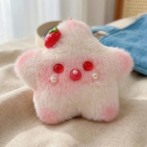 May include: A plush, star-shaped keychain in shades of pink and white, with a red strawberry accent. The keychain features red gem eyes and a pink gem nose, with pearl accents. The plush toy is soft and fluffy.