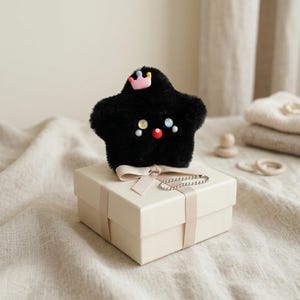 May include: A black plush star-shaped toy with a pink crown and colorful accents, resting on a cream-colored gift box tied with a ribbon. A silver chain is draped over the box. The toy has a cute face with colorful embellishments.