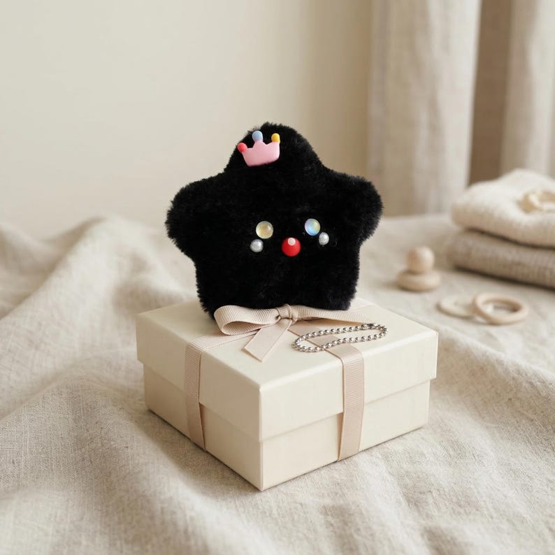 May include: A black plush star-shaped toy with a pink crown and colorful accents, resting on a cream-colored gift box tied with a ribbon. A silver chain is draped over the box. The toy has a cute face with colorful embellishments.