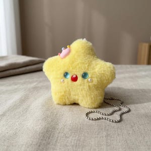 May include: A plush yellow star-shaped keychain with blue, red, and pearl accents. The star has a small pink crown and a silver chain. The soft toy is on a neutral-colored surface.