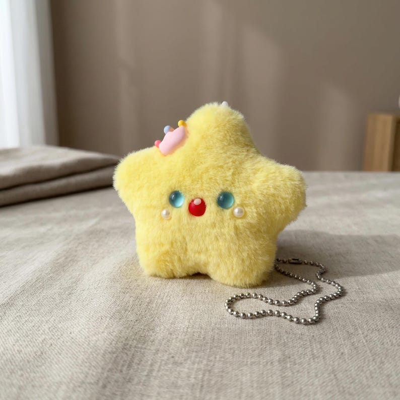 May include: A plush yellow star-shaped keychain with blue, red, and pearl accents. The star has a small pink crown and a silver chain. The soft toy is on a neutral-colored surface.