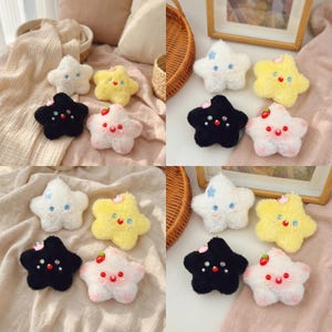 May include: A collection of plush star-shaped ornaments in various colors, including white, yellow, black, and pink. Each star features embroidered details such as eyes, a mouth, and decorative accents. Some stars have a small strawberry or bow.