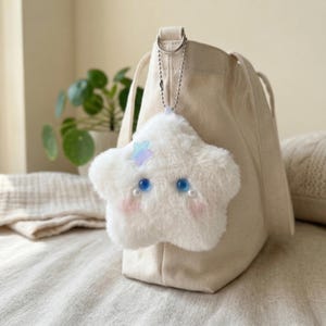 May include: A fluffy white star-shaped keychain with blue eyes, pink cheeks, and a small iridescent star accent. The keychain is attached to a beige tote bag with a silver chain. The bag is made of canvas-like material.