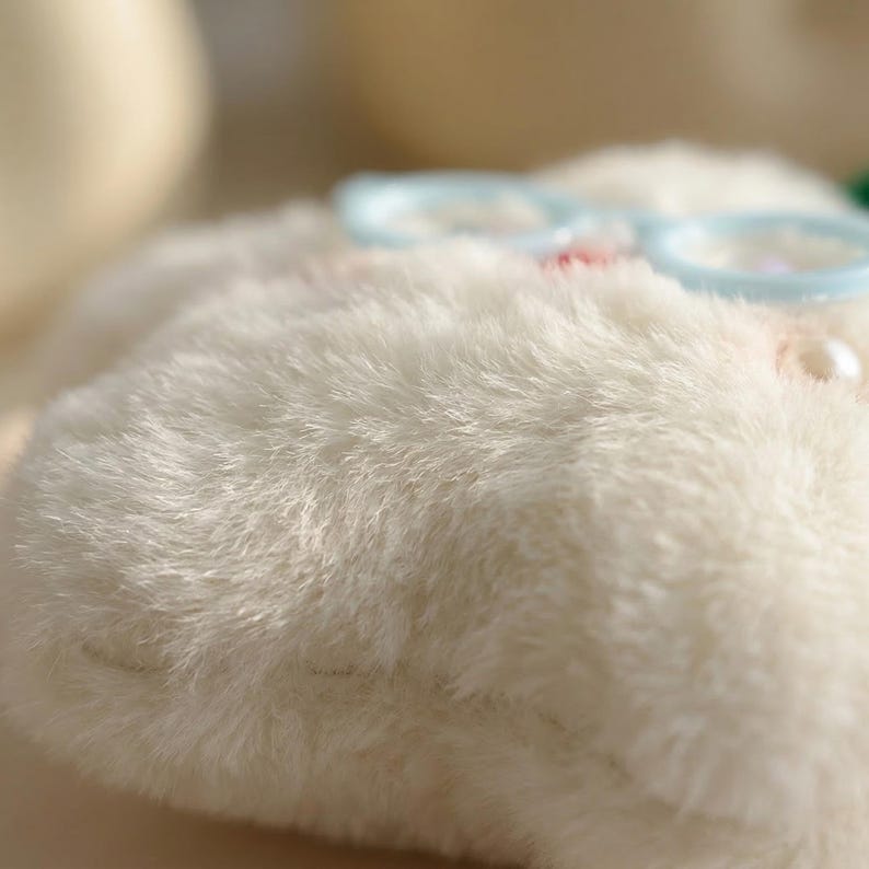 May include: Close-up of a fluffy, off-white plush item, possibly a toy or decorative piece. Light blue, round-framed glasses rest on top, with a small pearl accent. The plush material has a soft, textured appearance.