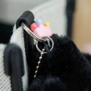 May include: Close-up of a black fluffy keychain with a pink crown charm and a silver chain. The keychain is attached to a black handle and a white and black patterned fabric.