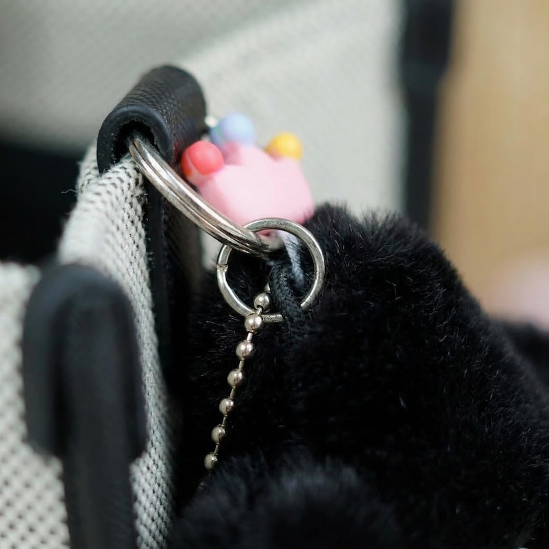 May include: Close-up of a black fluffy keychain with a pink crown charm and a silver chain. The keychain is attached to a black handle and a white and black patterned fabric.