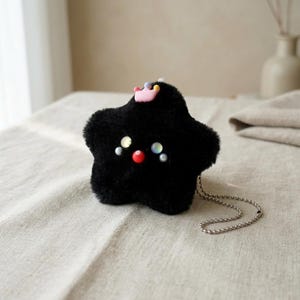May include: A black plush star-shaped keychain with a small pink crown and beaded chain. The star has a face with iridescent and red accents. The plush toy is on a neutral-colored surface.