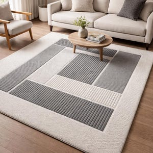 May include: A rectangular area rug with a geometric design. The rug features a cream-colored background with sections of gray and white rectangles and stripes. The rug is placed on a hardwood floor.