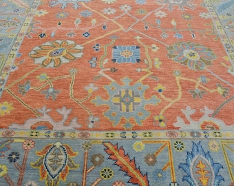 May include: A detailed close-up of a large area rug with an intricate floral design. The rug features a central field of orange-red with blue and yellow floral patterns. The border is a light blue with more floral motifs.