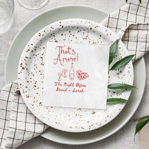 Pode incluir: Um guardanapo de papel branco com a frase "That's Amore!" em vermelho, juntamente com ilustra&ccedil;&otilde;es de uma garrafa de vinho, ta&ccedil;as e uma pizza em forma de cora&ccedil;&atilde;o. O texto "The Night Before David + Sarah" tamb&eacute;m est&aacute; presente. O guardanapo est&aacute; sobre um prato.