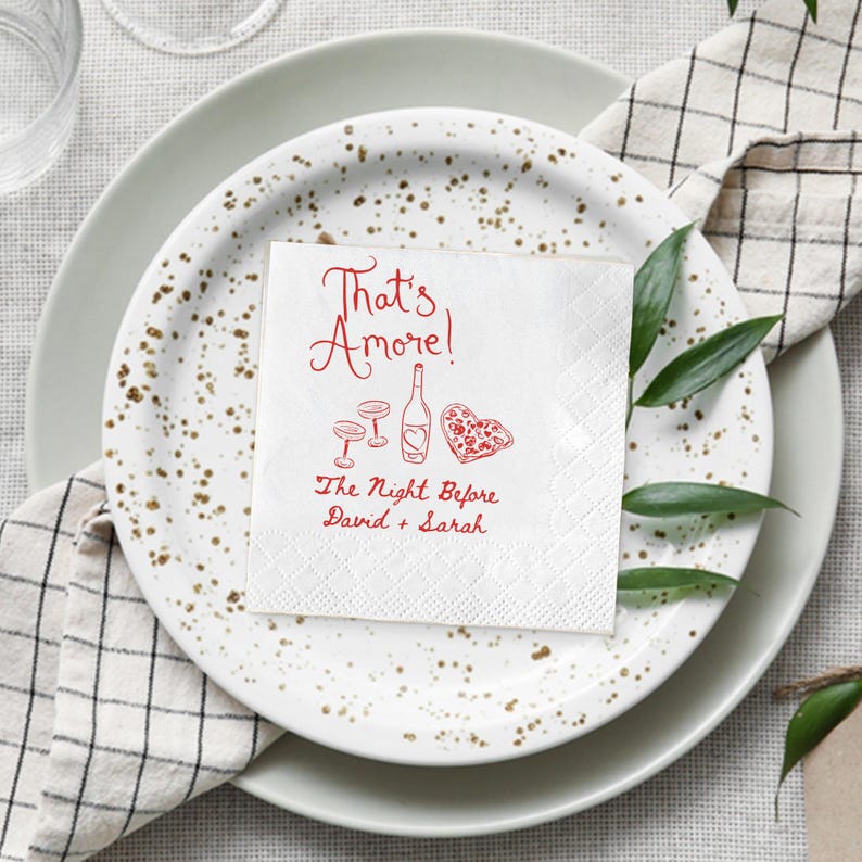 Pode incluir: Um guardanapo de papel branco com a frase "That's Amore!" em vermelho, juntamente com ilustra&ccedil;&otilde;es de uma garrafa de vinho, ta&ccedil;as e uma pizza em forma de cora&ccedil;&atilde;o. O texto "The Night Before David + Sarah" tamb&eacute;m est&aacute; presente. O guardanapo est&aacute; sobre um prato.