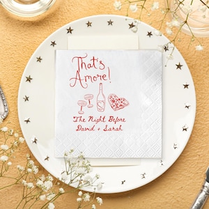 Pode incluir: Um guardanapo de papel branco com o texto "That's Amore!" em escrita vermelha, juntamente com ilustra&ccedil;&otilde;es de uma garrafa de vinho, dois copos e uma pizza em forma de cora&ccedil;&atilde;o. O texto "The Night Before David + Sarah" tamb&eacute;m est&aacute; presente.