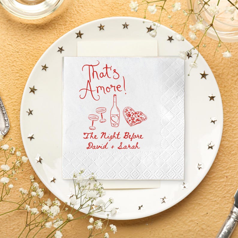 Pode incluir: Um guardanapo de papel branco com o texto "That's Amore!" em escrita vermelha, juntamente com ilustra&ccedil;&otilde;es de uma garrafa de vinho, dois copos e uma pizza em forma de cora&ccedil;&atilde;o. O texto "The Night Before David + Sarah" tamb&eacute;m est&aacute; presente.