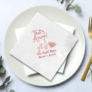 Pode incluir: Guardanapo quadrado branco com padr&atilde;o de losangos, com texto e ilustra&ccedil;&otilde;es vermelhas. O texto diz "That's Amore!" com imagens de ta&ccedil;as de vinho, uma garrafa e uma pizza em forma de cora&ccedil;&atilde;o. Abaixo, diz "The Night Before David + Sarah".