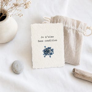 Peut inclure: Une carte texturée blanc cassé avec le texte français "Je m'aime Sans condition" et un motif floral bleu. Un petit sac à cordon beige et une pierre décorative sont à côté de la carte. Un petit vase avec des fleurs séchées est en arrière-plan.