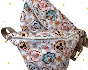 May include: Two beige fanny packs with a repeating pattern of Mickey Mouse, Donald Duck, and other cartoon characters in flower-shaped designs. The bags have gold zippers and adjustable straps.