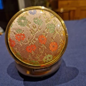 Puede incluir: Un estuche compacto redondo y vintage con un marco de metal dorado y una parte superior de tela floral. La tela presenta un patrón de flores rojas, verdes y azules sobre un fondo beige. El estuche compacto tiene una base plateada.
