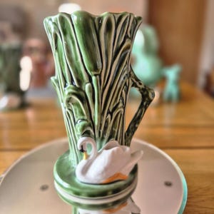 May include: A green ceramic vase with a swan design. The vase features a textured cattail pattern and a white swan with an orange beak. The vase has a handle and sits on a round, reflective surface.