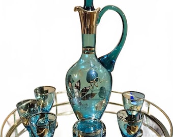 May include: A vintage teal glass decanter set with gold accents, featuring a decanter with a handle and stopper, and six matching glasses. The set is decorated with gold floral designs and sits on a mirrored tray.