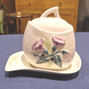 Op de afbeelding: Een witte keramische suikerpot met een deksel en een bijpassende schotel. De kom is versierd met paarse windebloemen en groene bladeren. De handgreep op het deksel is gebogen.