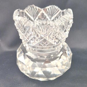 Puede incluir: Un portavelas de cristal tallado transparente con base facetada y borde festoneado. El intrincado diseño presenta patrones de diamantes y abanicos, creando un efecto brillante. Esta pieza decorativa es ideal para añadir un toque de elegancia.