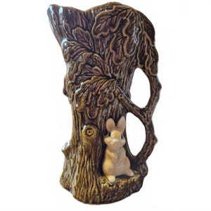 Könnte beinhalten: Braune Keramikvase in Form eines Baumstamms mit detaillierten Rinden- und Blattmustern. Eine kleine, hellfarbene Hasenfigur sitzt am Fuß. Die Vase hat eine Öffnung an der Oberseite für Blumen oder dekorative Zwecke.