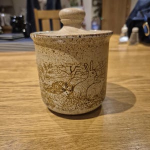 Op de afbeelding: Een beige keramische pot met een deksel, met een natuurscène met konijnen en een kip. De pot heeft een gespikkelde textuur en een afgeronde vorm. Het deksel heeft een kleine, ronde knop. De pot staat op een houten tafel.