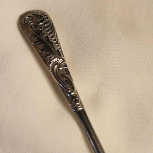 May include: Close-up of a silver spoon with ornate, engraved floral and leaf patterns on the handle. The spoon has a curved, oval-shaped bowl and a long, slender handle. The design is detailed and intricate.