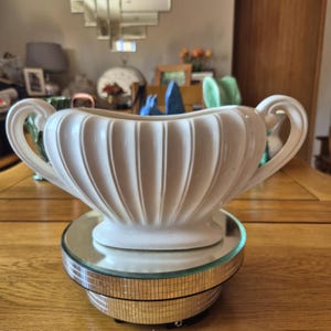 Peut inclure: Un pot de fleurs en céramique crème de style vintage, avec un design cannelé et des poignées incurvées. Le pot est présenté sur un présentoir rotatif miroir. L'arrière-plan comprend une table en bois et un miroir décoratif.