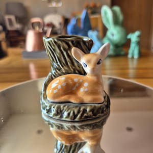 May include: A ceramic ornament featuring a fawn with white spots, set on a decorative, tree-trunk-shaped base. The fawn is light orange with white spots. The base is brown and green. Other ceramic ornaments are in the background.