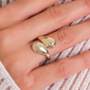 May include: A gold-colored ring with a unique design featuring two teardrop-shaped elements. The ring is worn on a hand, showcasing its polished finish and elegant style. The ring's design is modern and eye-catching.