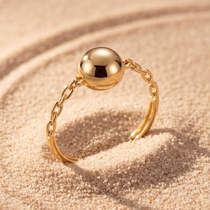May include: A gold-toned ring featuring a polished, spherical bead at the center. The band is constructed from delicate, interconnected metal chain links, creating a flexible and open design that rests on a textured, sandy surface.