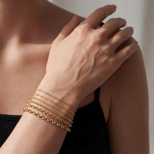 May include: A collection of gold bracelets of varying chain styles, including delicate cable chains and a thicker, rounded chain. The bracelets are stacked on a wrist, showcasing the different designs and textures of the jewelry.