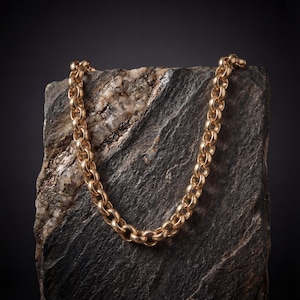 May include: A gold-colored chain necklace with a rounded link design. The necklace is displayed on a dark gray and tan stone surface against a black background. The chain is made of metal and has a polished finish.