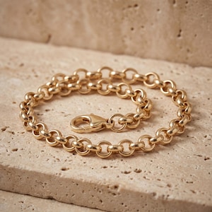May include: A gold-colored bracelet with a lobster clasp. The bracelet is made of a series of connected, rounded links. The jewelry is displayed on a light beige surface.