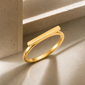Könnte beinhalten: Ein polierter goldfarbener Ring mit einem glatten, horizontalen Balkendesign auf der Oberseite. Das Band ist dünn und kreisförmig und verbindet sich mit den Enden des geraden, flachen Balkenelements. Die Metalloberfläche hat ein glänzendes Finish.