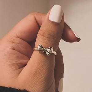 May include: A silver ring with a bow-shaped design. The ring is worn on a finger, highlighting the intricate details of the bow. The ring's band is slender, and the bow is the main feature of the jewellery.