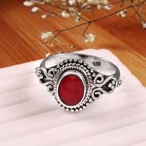 May include: A silver ring featuring an oval, deep red gemstone. The ring has an intricate design with beaded and swirling patterns around the central stone. It rests on a white surface, with a wooden backdrop and floral decorations.