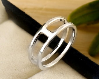 Könnte beinhalten: Ein silberner Ring mit einem einzigartigen offenen Design. Der Ring hat ein Doppelband mit einer rechteckigen Öffnung in der Mitte. Der Ring wird in einer schwarzen Schmuckschatulle mit beigem Innenraum präsentiert.