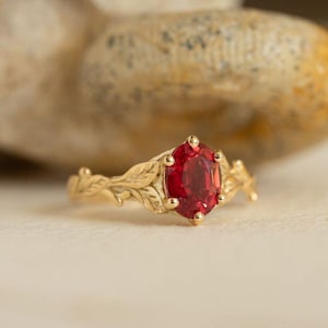 Puede incluir: Un anillo de oro con una gran gema roja de corte ovalado. La banda del anillo presenta un delicado diseño de hojas. La gema es el punto focal, engastada en un engaste de oro con pequeñas garras.