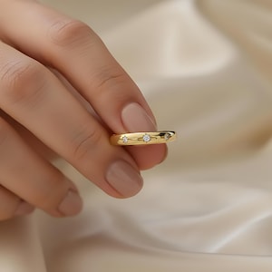 May include: A gold ring featuring three small, clear gemstones. The ring has a smooth, polished finish and a rounded band. The gemstones are set in a row. The ring is held in a hand. The gemstones sparkle against the gold.