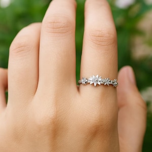 Puede incluir: Un anillo plateado con un diseño de estrellas. El anillo está adornado con pequeñas piedras brillantes, formando un patrón celestial. El anillo se lleva en un dedo, mostrando sus detalles delicados e intrincados.